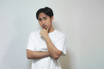 A young Asian man pondering, chin resting on hand, looking thoughtful.