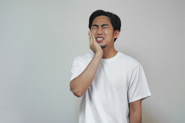Man in white shirt grimaces, holding his cheek in pain, possibly experiencing a toothache or facial...