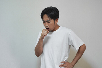 A man in a white t-shirt coughs into his hand, appearing unwell against a plain background.