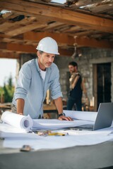 A portrait of an architect working on construction plans with a pen while standing at a table inside a new house under construction.