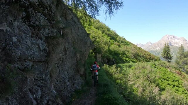 Two bikers hug cliffside singletrack, steep slope drops low, scattered pines frame distant horizon under bright clear sky.