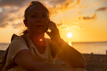 Woman talking on the phone during sunset at a beach, enjoying the serene atmosphere while the sun...