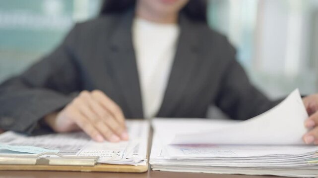 A woman is sitting at a desk with a stack of papers in front of her. She is holding a piece of paper and looking at it