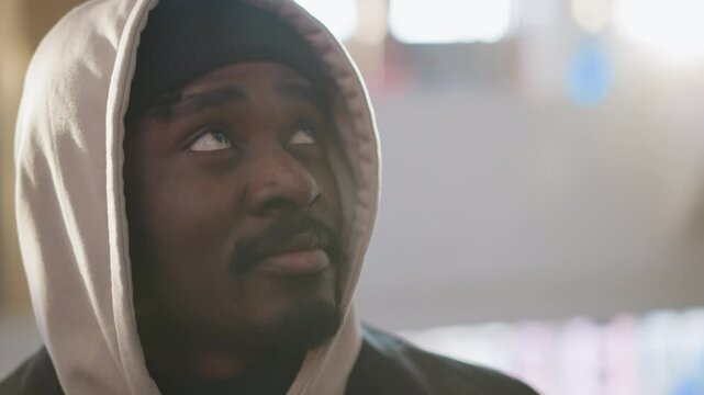 young black chap in beige hoodie and black beanie gazing upward inside sunlit modern interior space with blurred windows and reflective metal elements conveying curiosity and calm introspection