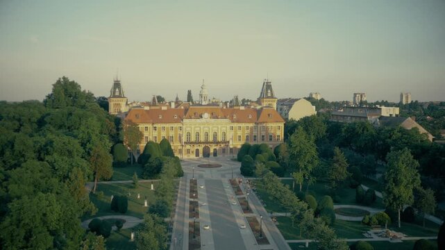 Majestic aerial pan of Sombor City Hall (The Zupanija) in Serbia. The shot captures the historic architecture and surrounding manicured park during a beautiful golden hour.
