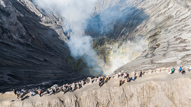 Aerial view of wisps of smoke rise from the Bromo volcano crater as a line of tiny figures traverse the steep, rocky path, Mount Bromo, East Java, Indonesia.