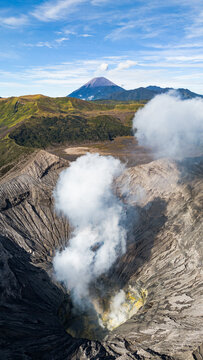 Aerial view of smoke billowing from the dark, textured crater of Mount Bromo, contrasting against the distant blue mountain, Mount Bromo Volcano, East Java, Indonesia.
