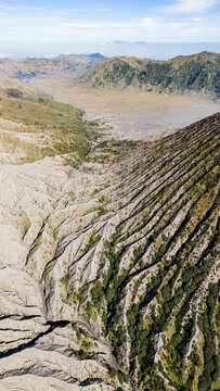 Aerial view of a rugged landscape where stark ridges meet a vast, dusty plain under a serene sky, Mount Bromo Volcano, East Java, Indonesia.