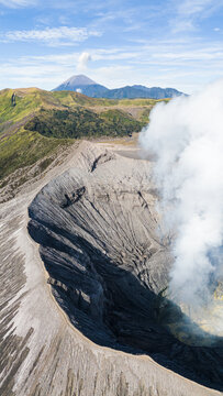 Aerial view of the rugged, volcanic landscape, with wisps of smoke rising from the crater, against a backdrop of distant peaks, Mount Bromo Volcano, East Java, Indonesia.