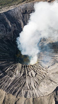 Aerial view of Mount Bromo's rugged caldera exhales a plume of ethereal white smoke against the dark, textured volcanic landscape, Mount Bromo Volcano, East Java, Indonesia.