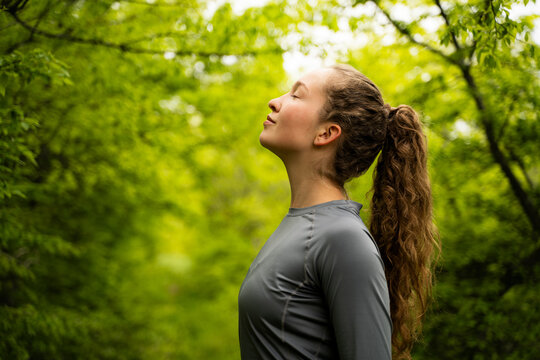 Young woman breathing fresh air in lush green forest practicing breath exercises enjoying peaceful moment in summer nature park wearing grey activewear, eyes closed, side view portrait