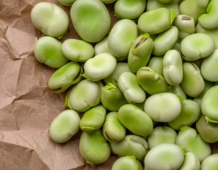 Close-up of a pile of fresh, green fava beans placed on a brown, crinkled paper surface. The beans...