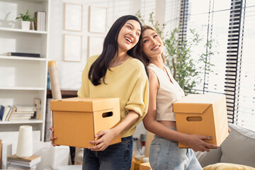 Happy Asian girl friends holding cardboard boxes moving to the new home. LGBT couple smiling, laughing with happiness for moving house together. Concept of buying and decorating property