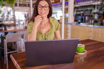 Female freelancer sipping cappuccino, working on laptop with wireless headphones at trendy cafe,...