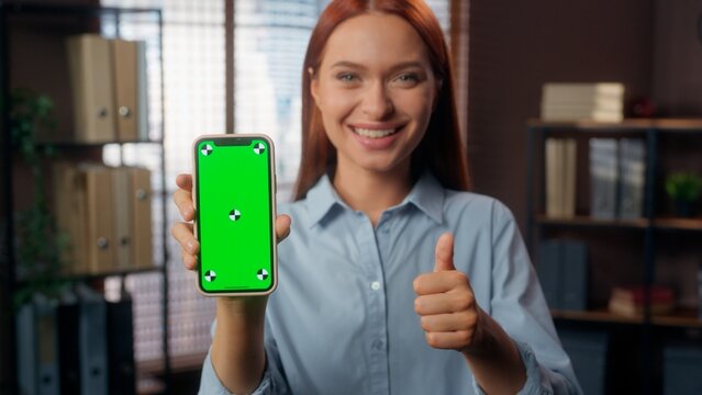 Happy smiling Caucasian business woman showing green screen mobile phone display smartphone app chroma key recommend show like thumb up approve e-commerce online female girl businesswoman in office