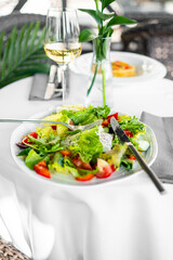 Fresh vegetable salad with tomatoes, cucumbers, lettuce, and feta cheese on a plate, served with white wine in an elegant dining setting.