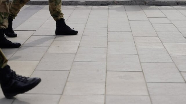 Two individuals in camouflage uniforms and black boots walk across a large paved open area, with one person exiting the frame, suggesting purposeful movement outdoors.
