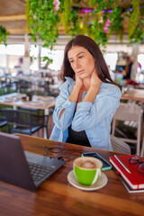 Woman seated at cafe, rubbing neck while typing on laptop, signaling workplace tension and...