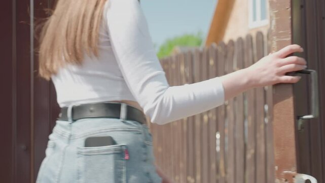 Rear view of lady wearing white crop top and jeans with smartphone in back pocket opening brown metal door as friend follows closely behind during sunny day, casual urban outdoor movement