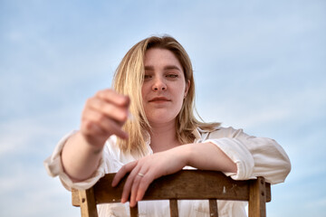 Young female artist extends paintbrush, inviting creativity. Set outdoors against serene blue sky,...