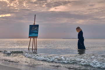 Young woman stands beside easel on serene beach, with calm sea and overcast skies creating...