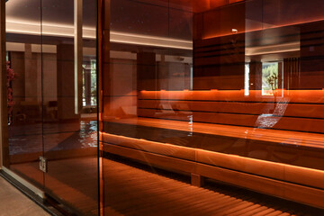 Modern wooden sauna with warm lighting behind a glass wall, located near an indoor pool with water...