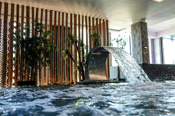 Indoor spa pool with a curved metal waterfall, tropical plants, and lounge chairs. Elegant modern...