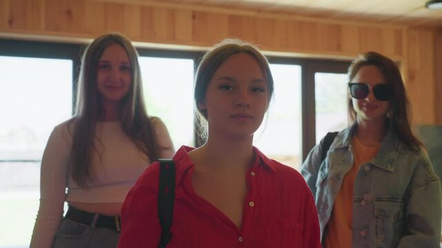 Three young girls pose indoors near window in wooden cabin as lady in sunglasses begins to remove them with confident expression, warm light creating cozy mood and stylish casual atmosphere