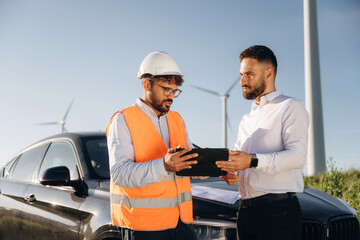 Instructions for guy in hard hat. Two men engineers are on field, with wind turbines