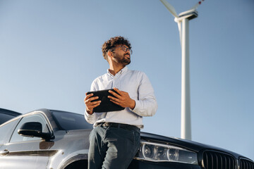 By the front of the car, with tablet. Male engineer in field checking on wind turbine production