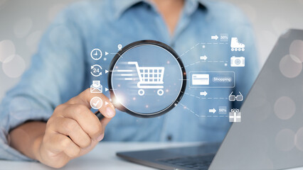 Hand holding a magnifying glass focusing on a shopping cart icon with various online shopping and e-commerce icons, representing digital shopping, product search, and e-commerce marketing.