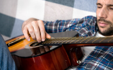 Close-up of a young man strumming an acoustic guitar while relaxing at home. Concept of music hobby, home practice, creativity, and relaxation.