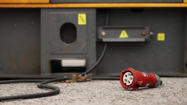 Industrial power connector unplugged from electrical equipment with visible warning labels and cables placed on rough outdoor ground surface in an operational setting