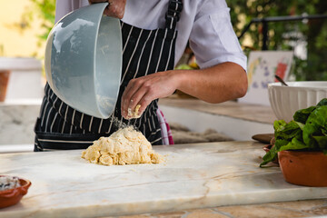 A chef in a striped apron pouring dough from a ceramic mixing bowl onto a floured countertop in a...