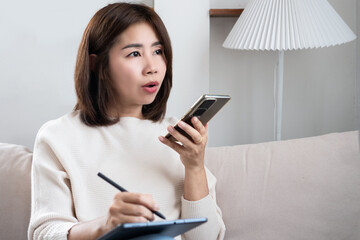 Asian woman talking to a voice assistant on her smartphone while working from home, using AI chatbot technology to get helpful ideas and writing them down on a tablet