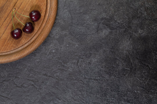 Dark Sweet Cherries on Round Wooden Cutting Board with Slate Background. In upper left corner. The rest of the frame leaves plenty of empty space for text or design.
