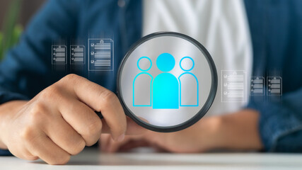 Hand holding magnifying glass focusing on people icon with resume documents, representing recruitment, human resources, candidate screening, and talent acquisition.