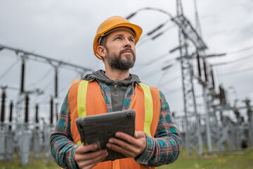 Obraz premium An electrical engineer inspecting a power substation in the field, wearing a hard hat and reflective vest, holding a tablet, high-voltage towers in the background