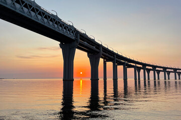 Beautiful sunset over the sea. The sun setting between the pillars of the bridge. Reflection of sunlight on water. The sky is painted in golden colors. Kanonersky Island, Saint-Petersburg, Russia.