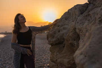 Fototapeta premium A young Caucasian woman with long brown hair stands by a rocky beach at sunset. She wears a black top and maroon pants, enjoying the serene atmosphere.