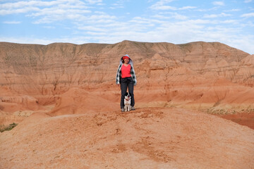 A happy woman smiles, she stands against the backdrop of the beautiful red mountains of Boguta, her dog is next to her, they travel together through the unique nature of Kazakhstan.