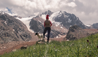 Rear view of woman in hiking clothes, down vest and red hat against mountains background, she is hiking with her dog looking at impressive snow capped mountain peaks