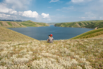 A woman and her dog friend sit in a field of mountain edelweiss on the shore of a lake in summer, admiring the surrounding landscape of the wild nature of Kazakhstan. Bestyubinskoye Reservoir