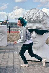 Young woman with blue hair and denim jacket run happily near urban artwork. Bright sunlight. Urban...