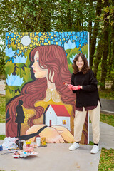 Young female with wavy brown hair standing by colorful en plein air canvas. Smiling, holding...