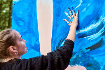 Young woman with blonde hair and joyful expression paints blue strokes on canvas using hands....