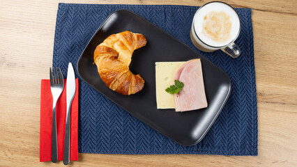 Top view of a breakfast croissant served with ham and cheese on a black plate, accompanied by a cappuccino on a wooden table with blue placemat. Classic savory European morning meal.