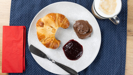 Overhead view of a breakfast croissant served with chocolate spread and berry jam on a white plate, sprinkled with powdered sugar, next to a cappuccino. Sweet and indulgent morning treat.