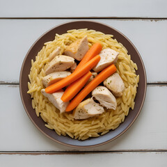 A plate of orzo pasta with chicken pieces and carrots on a white wooden surface from a top down view
