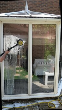 Vertical video of a mature nan using a power washer brush to clean the outside of a sunroom and windows with soapy water